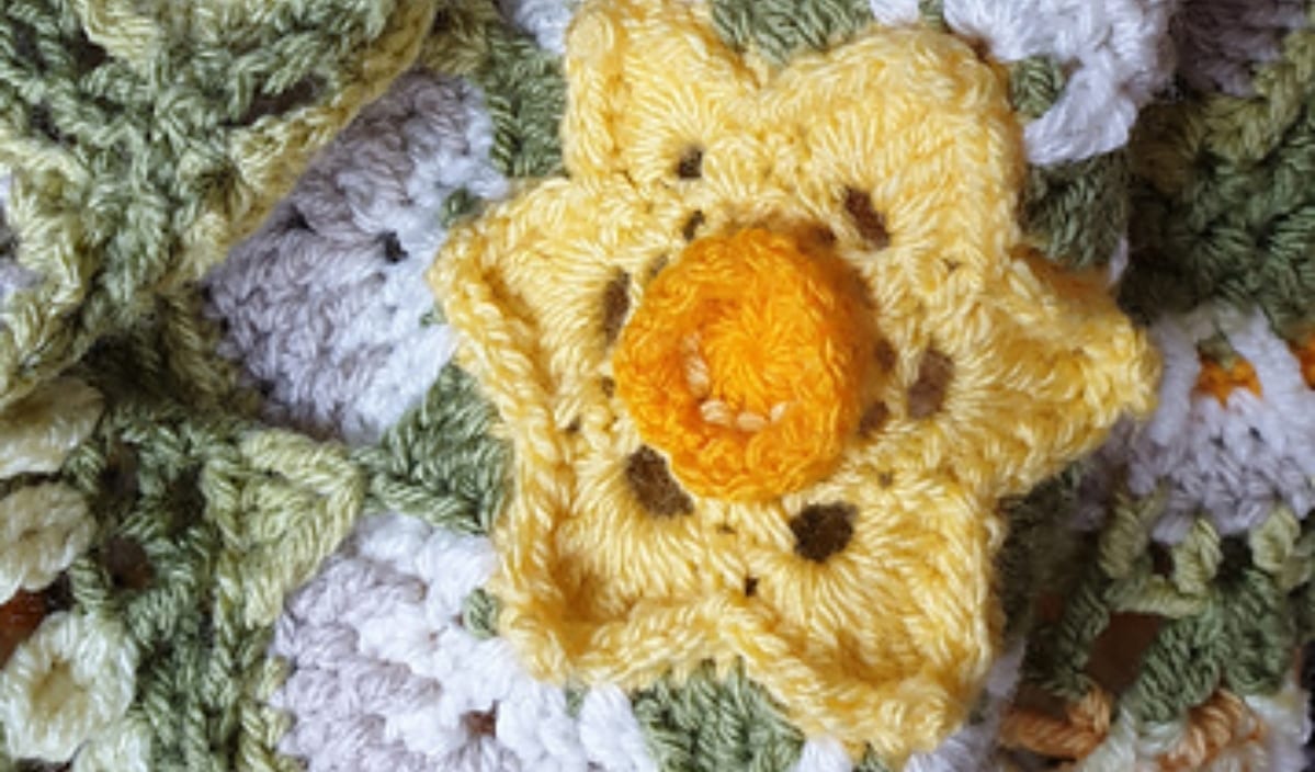 Close-up of a crocheted yellow and orange flower, resembling a 3D Flower Blanket design, with green and white yarn in the background.