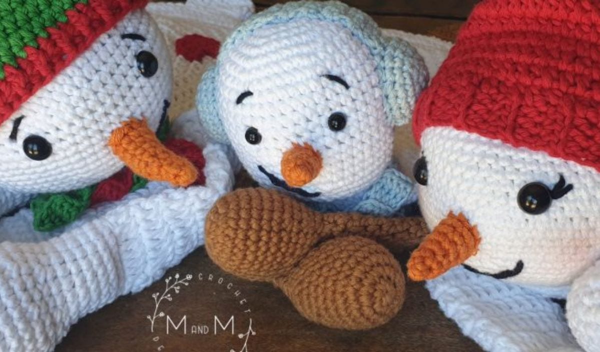 Three charming crocheted snowmen with carrot noses and hats lie on a wooden surface, ready to bring festive cheer. Their delightful design makes for perfect Christmas decor or an inspiration for your next crochet pattern project.