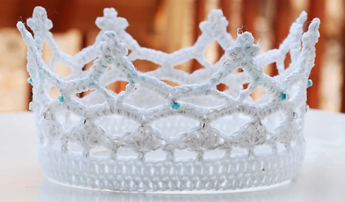 A cute crown, crafted with intricate white crochet patterns and adorned with small blue beads, rests elegantly on a light surface.