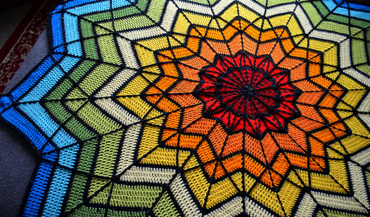 A colorful crochet blanket showcases a geometric, starburst pattern with red, orange, yellow, green, and blue sections outlined in black, reminiscent of a mosaic square.