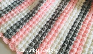 A crocheted blanket, aptly named the Sweet Dream Blanket, showcases alternating stripes of pink, white, and gray yarn in a textured pattern. The text "Lullaby Lodge" graces the corner, perfect for those seeking free crochet patterns.