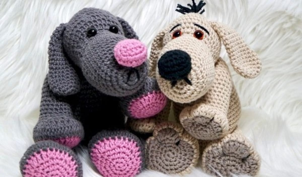 Two crocheted stuffed dogs, crafted from free crochet patterns, sit side by side on a white furry surface. One is gray with pink accents, and the other is beige with a black nose.