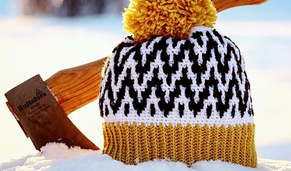 A winter hat with yellow, black, and white patterns and a pom-pom sits on snow alongside a wooden-handled axe, showcasing intricate crochet patterns.