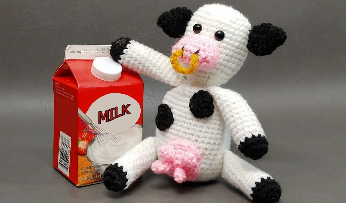 A crocheted cow doll, crafted from cute cow crochet patterns, sits beside a red milk carton against a plain gray background.