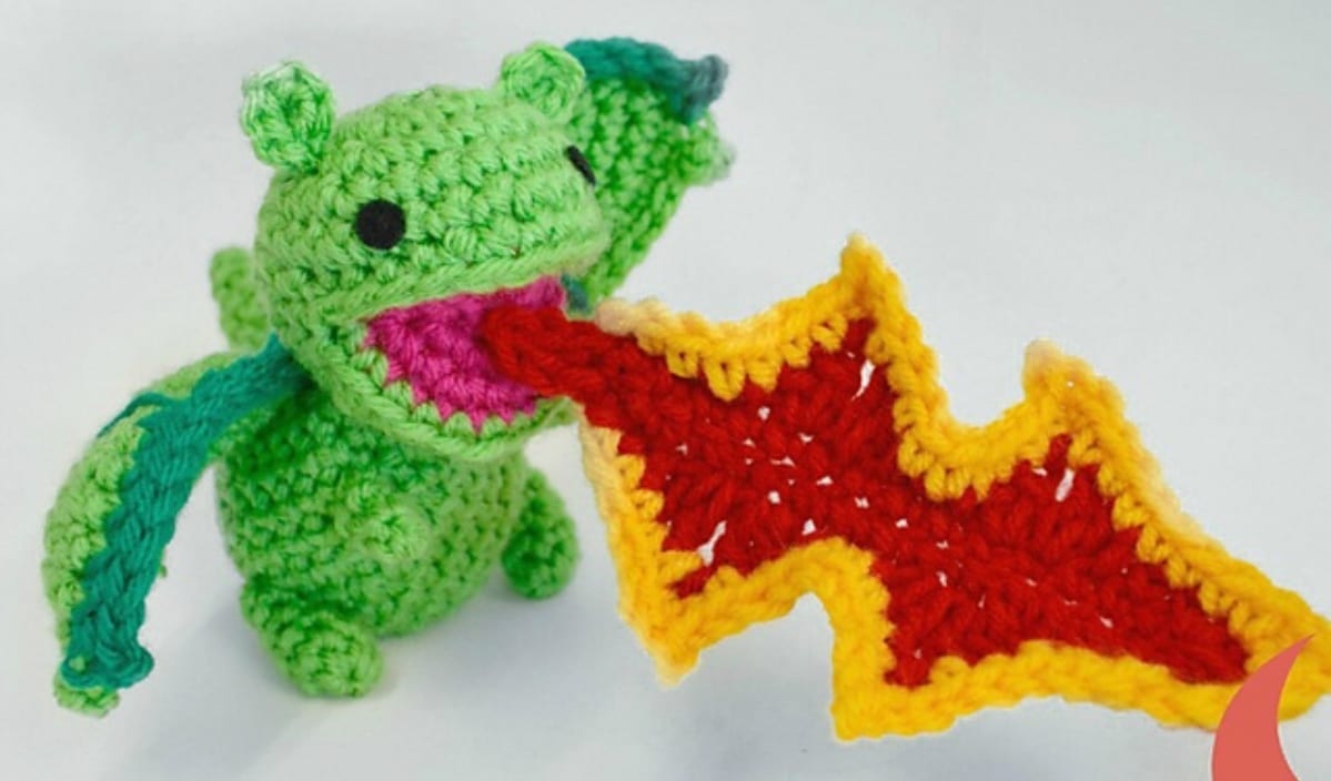 A crocheted green dragon with blue wings breathes a bright red and yellow crocheted flame, inspired by whimsical crochet patterns.