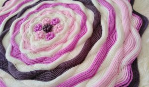 A round crochet baby blanket with concentric flower patterns in shades of pink, purple, and white.