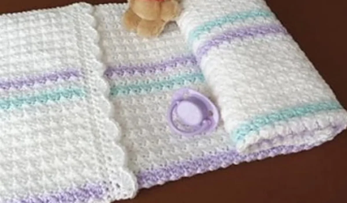 Crocheted baby blanket with scalloped edges in white, purple, and blue stripes, a purple pacifier, and a small teddy bear on a brown surface. Perfect for those seeking inspiration from free crochet patterns.