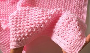 Hands holding a textured, light pink knitted fabric reminiscent of easy blanket crochet patterns.