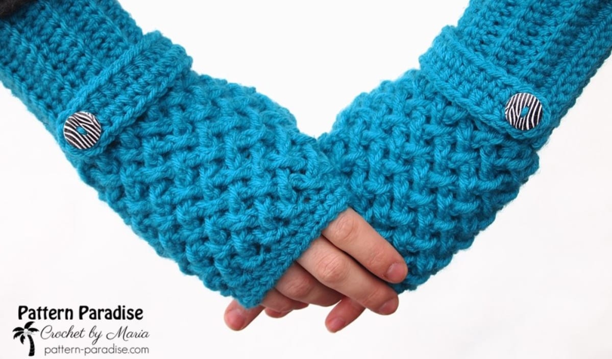 Close-up of hands wearing blue crocheted fingerless gloves with striped buttons, reminiscent of a dragon's scales. Logo: "Pattern Paradise.