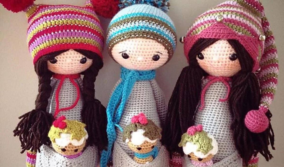 Three amigurumi dolls in colorful hats and scarves, each holding a small, knitted cupcake.