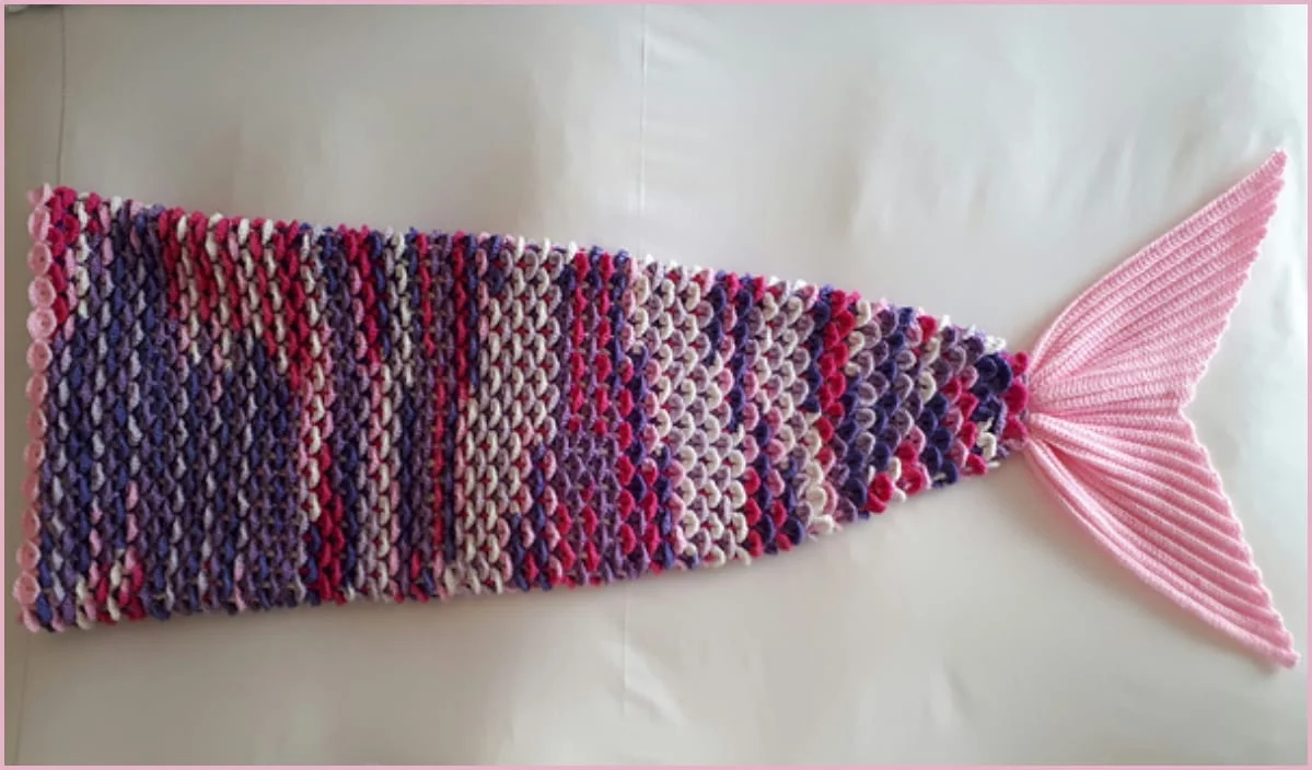 A crocheted mermaid tail blanket, crafted from a delightful mosaic blanket design with free crochet patterns, features multicolored scales and a pink tail fin, beautifully laid out on a white surface.