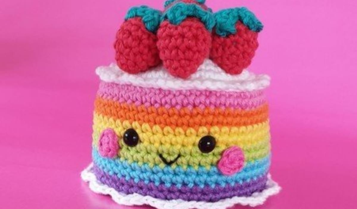 Colorful crocheted cake with a smiling face, featuring a rainbow pattern and topped with red strawberries, cleverly masquerades as an adorable crochet purse against a pink background.