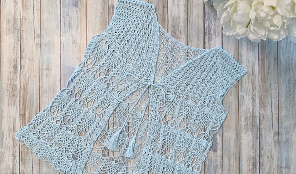 An airy summer vibe graces this light blue crochet vest, elegantly tied at the front and displayed on a wooden surface next to delicate white flowers, showcasing intricate crochet patterns.