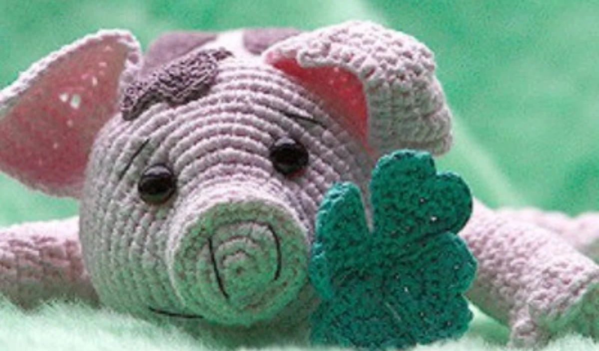 This crocheted mini pig with dark eyes and floppy ears holds a green clover against a soft green background. Perfect for those who love free crochet patterns and enjoy crafting adorable creations.