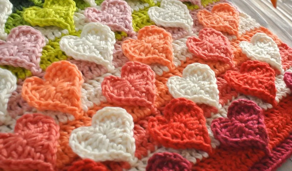 A beautiful crocheted blanket, reminiscent of a delicate doily, showcases rows of multicolored hearts in vibrant shades of pink, red, and cream.