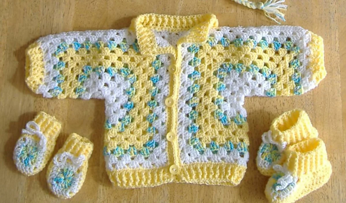 Crocheted yellow and white baby jacket with multicolored accents, alongside matching booties and mittens, laid on a wooden surface. Discover free crochet patterns to create your own adorable baby jacket set.