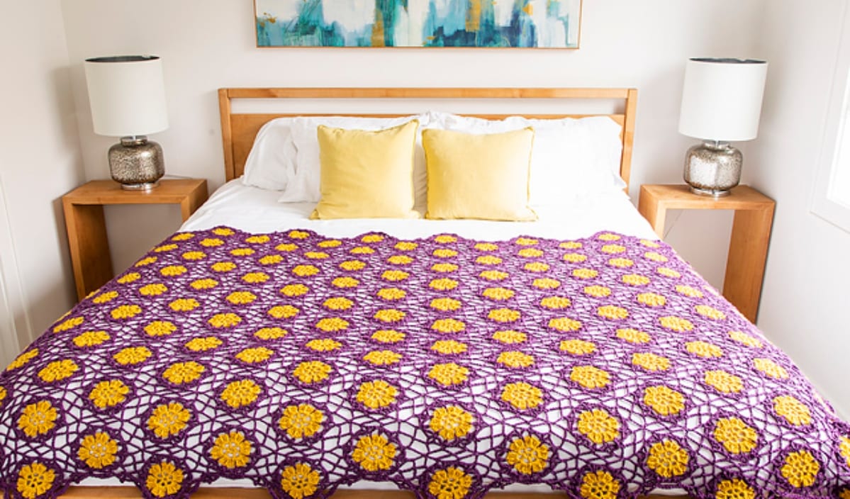 A bedroom featuring a wooden bed with white bedding, yellow pillows, and a purple and yellow crocheted blanket crafted from lovely lacy decor. Two wooden nightstands with lamps flank the bed, all under the serene gaze of abstract art on the wall.