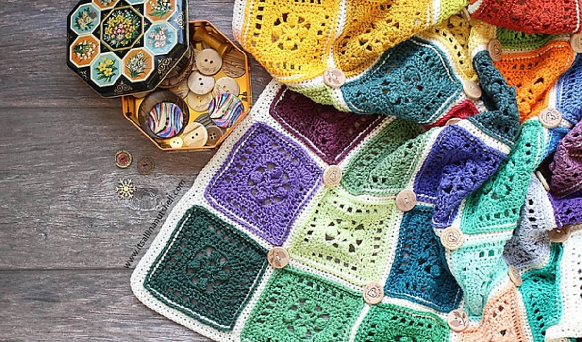 A colorful square blanket with crocheted patterns lies next to an open hexagonal box filled with assorted buttons on a wooden surface.