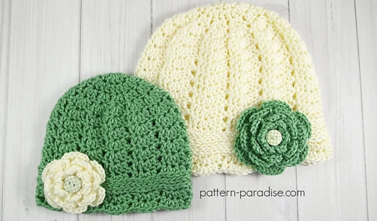 Two crocheted hats rest on a white surface: one green with a cute white flower and the other cream with a charming green flower. Both flowers have button centers. Perfect for summer, these hats could be made using free crochet patterns to add to your collection.