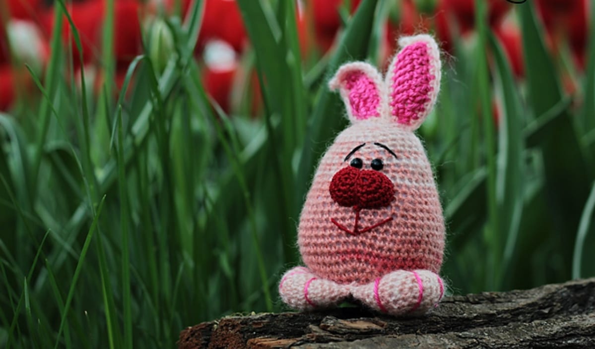 A crochet pink bunny with a red nose, inspired by classic Easter Bunny themes, sits on a log surrounded by tall grass.