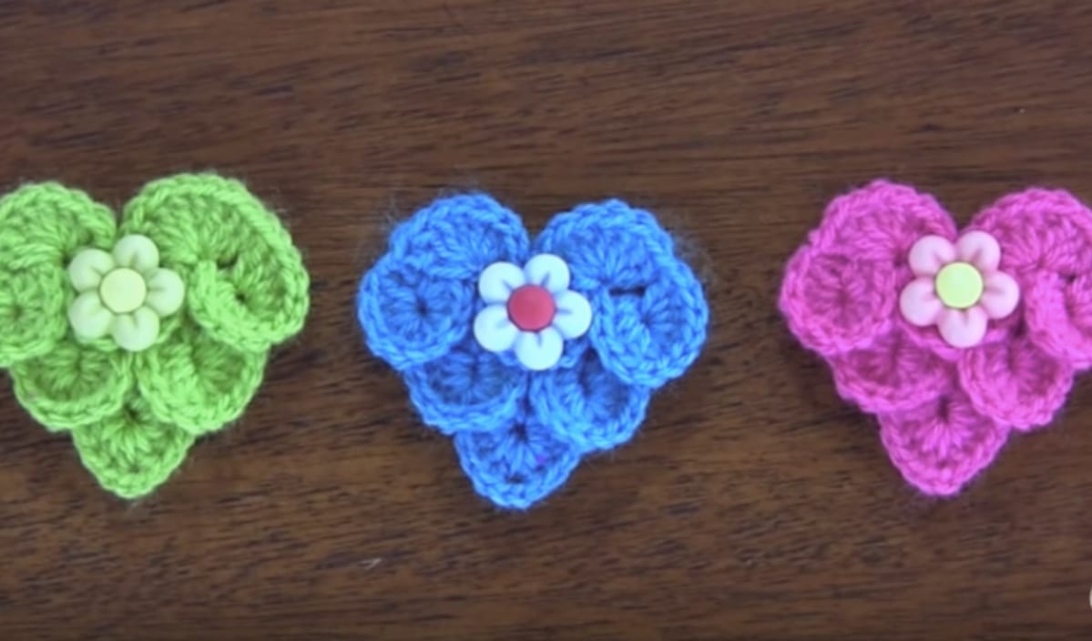 Three colorful crochet heart shapes in green, blue, and pink, each adorned with a white flower-shaped button at the center, are displayed on a wooden surface. Perfect for Valentine's patterns, these charming pieces add a touch of handmade love to any setting.