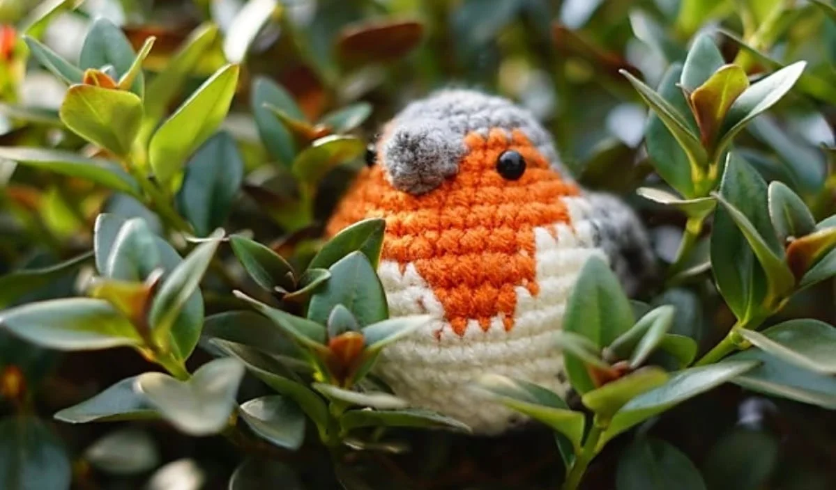 A knitted toy bird with a realistic orange breast is nestled among green leaves.