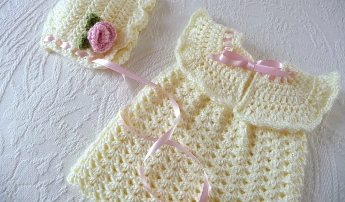 A crocheted baby dress and bonnet set in light yellow with pink ribbon accents and a pink flower detail on the bonnet, laid out on a textured white surface. Perfect for spring, this delightful Spring Baby Set is inspired by free baby crochet patterns.