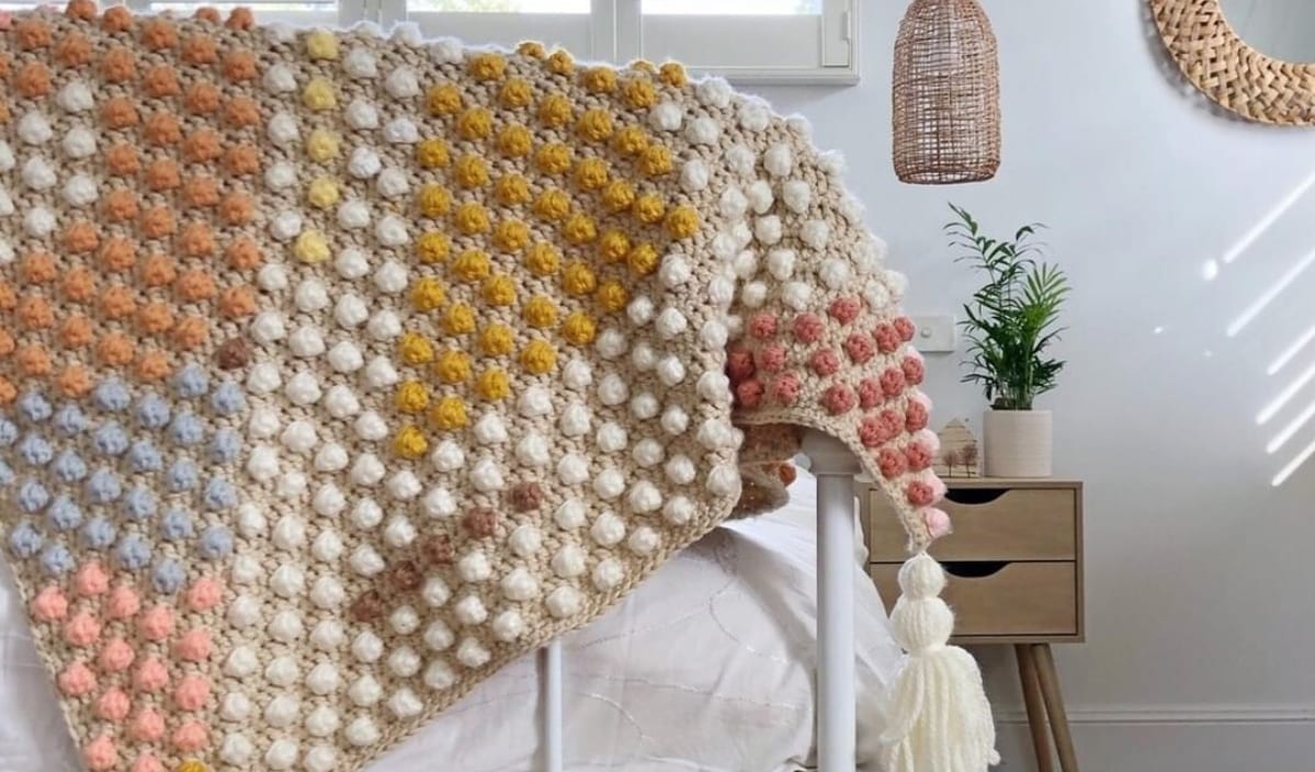 A textured, multicolored bobble blanket draped over the corner of a bed in a well-lit room with a wooden nightstand and potted fern beside it showcases exquisite crochet patterns.