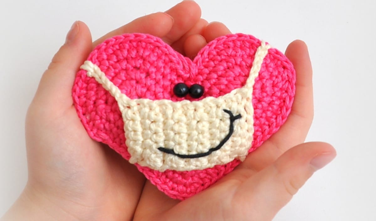 Hands cradling a pink crocheted heart with a smiling face and a cream-colored mask, crafted from free crochet patterns.