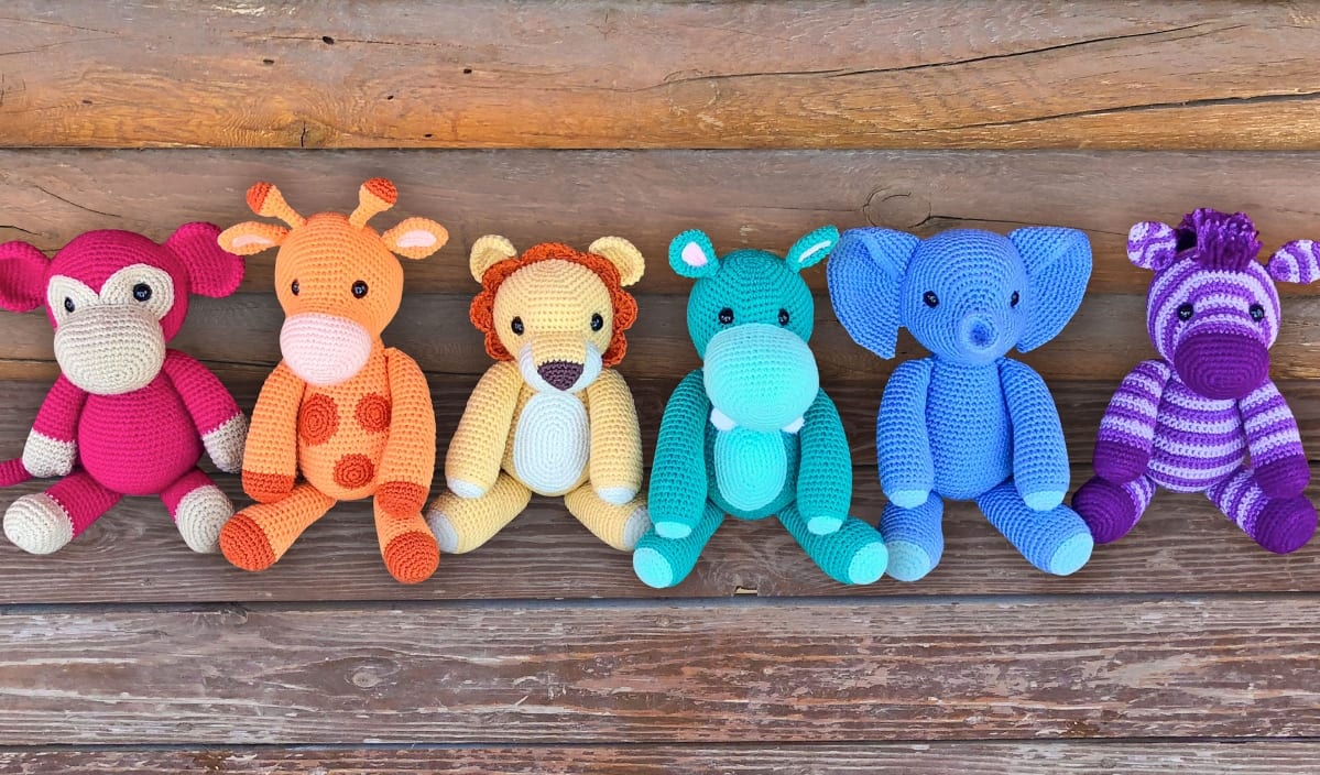 Six colorful crocheted animal dolls, perfect for a playful safari trip, are lined up on a wooden surface. They include a monkey, giraffe, lion, dinosaur, elephant, and zebra, each in a different vibrant color.