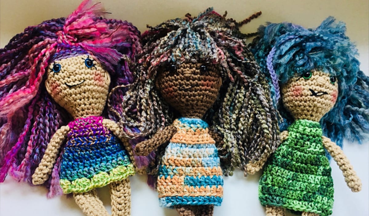 Three delightful crochet toys with colorful yarn hair and striped dresses, each featuring a cheerful expression, laid side by side.