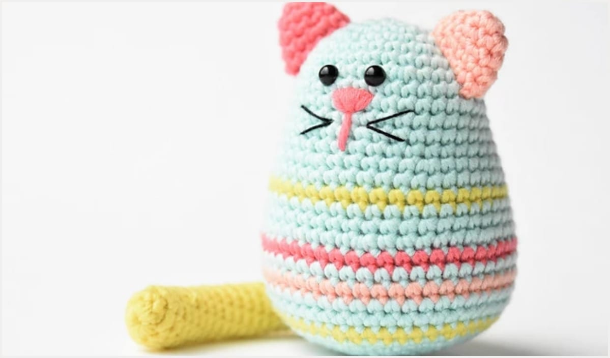 A crocheted toy resembling a cat with a striped body in pastel colors, featuring pink ears, black eyes, and a yellow tail. This adorable Ami comes to life using free crochet patterns, making it the perfect Easter egg surprise for any craft lover.