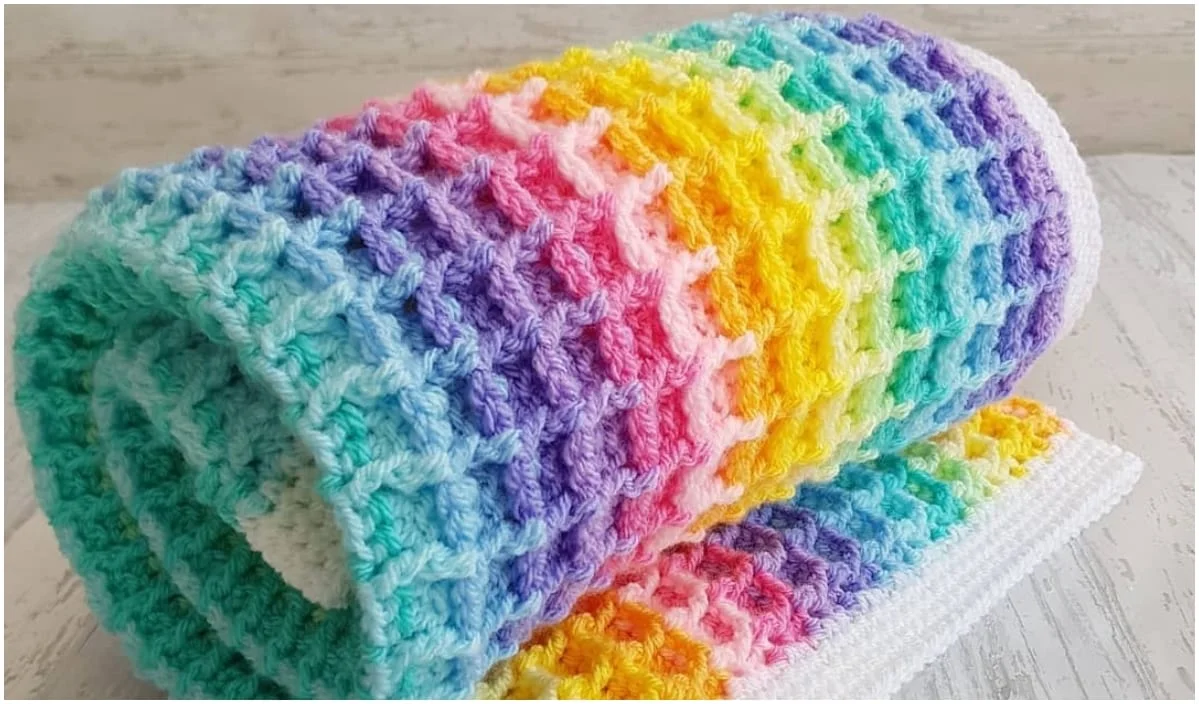 A vibrant waffle blanket, featuring a rolled, multicolored crochet design, is gracefully displayed on a light surface.