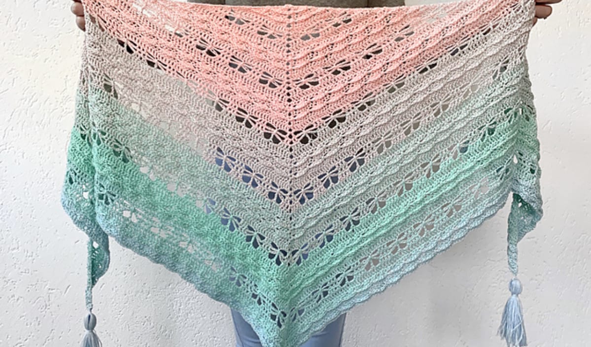 A person holds a Butterfly Shawl, a triangular crochet masterpiece with a gradient from pink to green. It features intricate lace patterns and tassels at the corners, beautifully crafted using free crochet patterns.