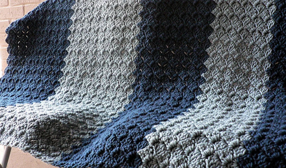 A crocheted blanket with alternating stripes of dark blue, medium blue, and light gray draped over a surface, reminiscent of soothing amigurumi patterns.