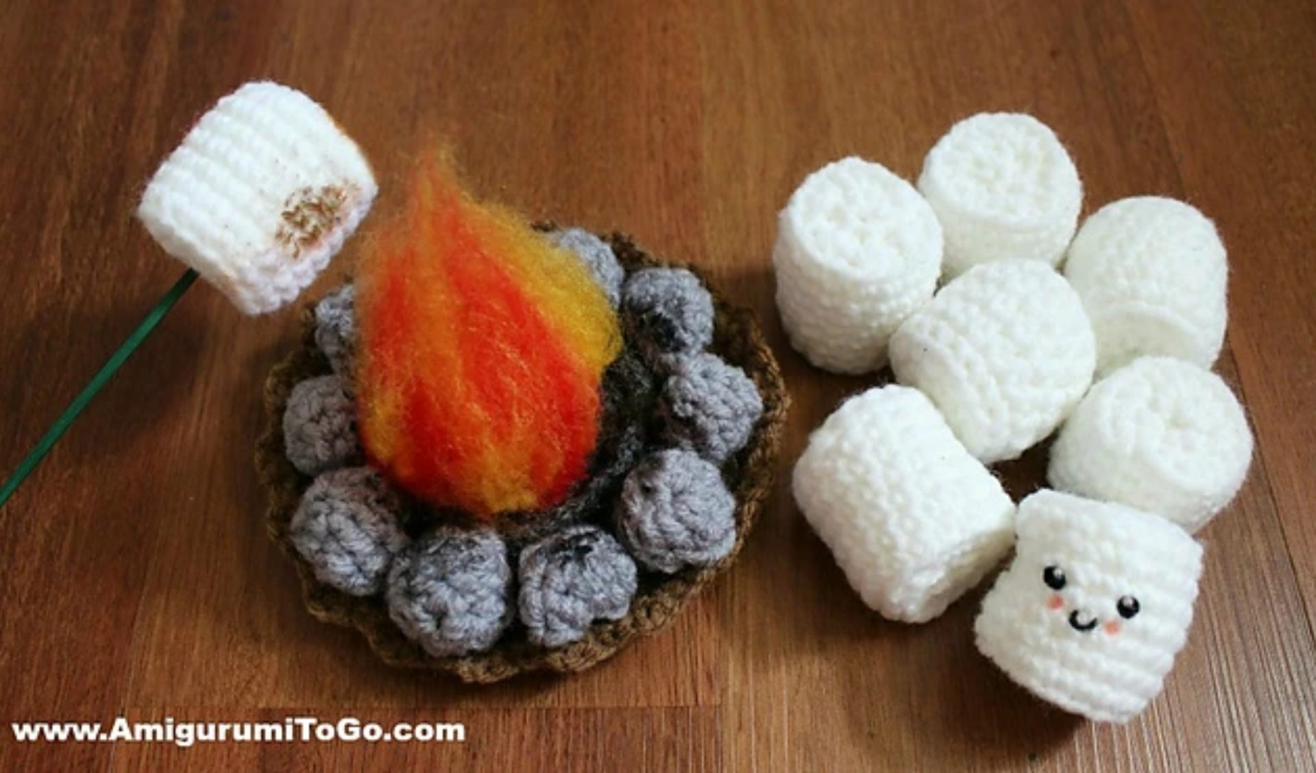 A delightful amigurumi campfire features three charming, smiling marshmallows with a skewer playfully toasting one atop a wooden surface.