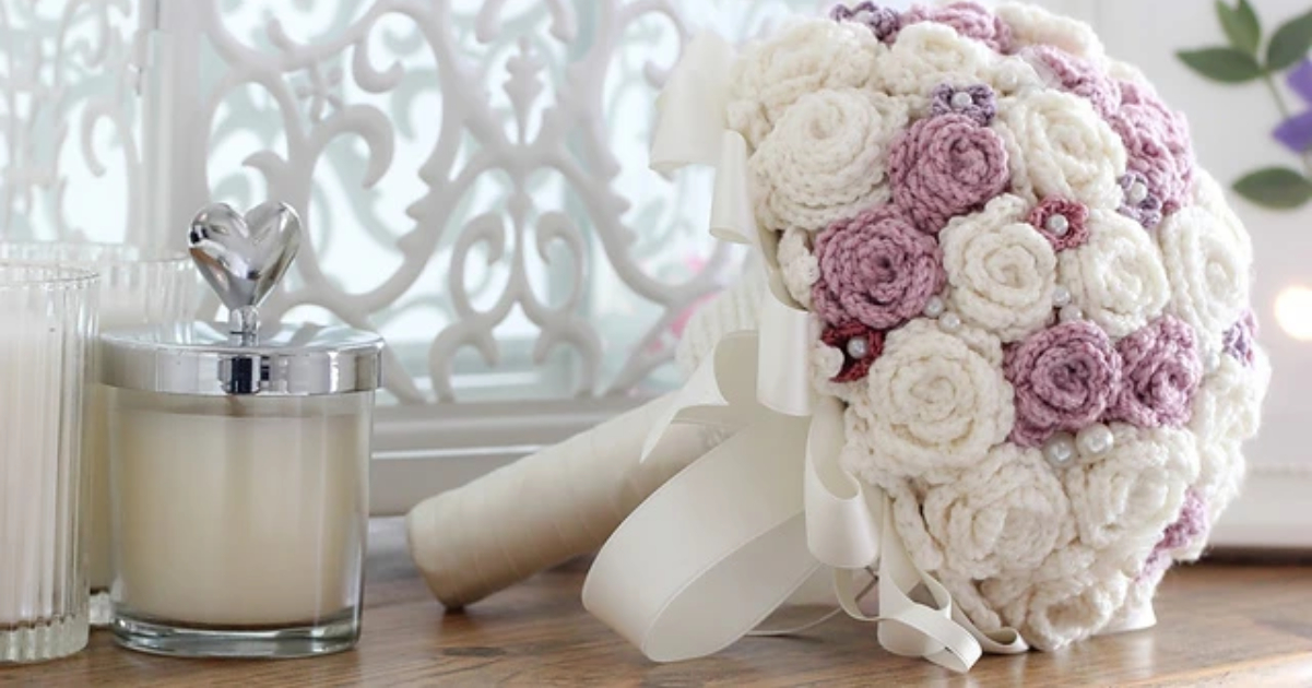 A crocheted bouquet with white and pink flowers, including delicate crocheted roses adorned with pearls, lies on a wooden surface next to a heart-topped candle.