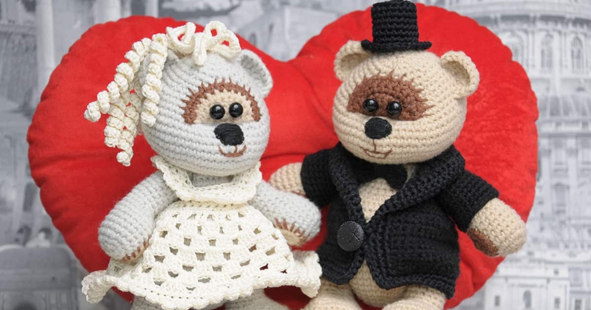 Two charming amigurumi teddy bears, dressed as a new couple ready for their wedding day, sit adorably in front of a large red heart backdrop. The bride wears a white dress, while the groom dons a black suit.