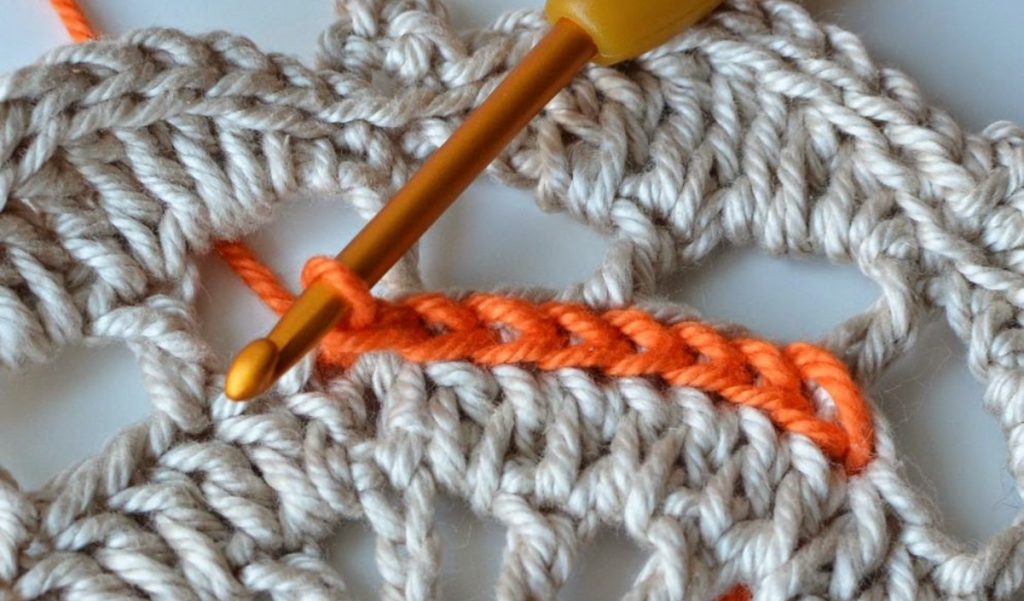 Close-up of a crochet project with a yellow hook and beige yarn, showcasing a surface crochet technique with an orange strand artfully woven through the pattern.