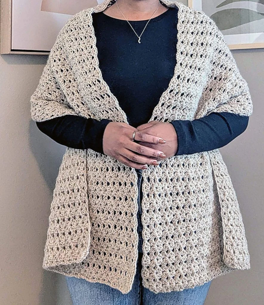 Person wearing a beige prayer shawl crochet pattern over a black long-sleeve shirt and blue jeans, standing indoors with hands clasped.