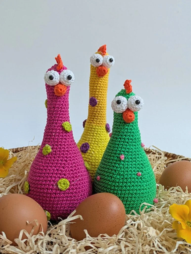 Three colorful crocheted chickens made from Easter crochet patterns with googly eyes stand on straw, surrounded by brown eggs and yellow flower petals against a white background.