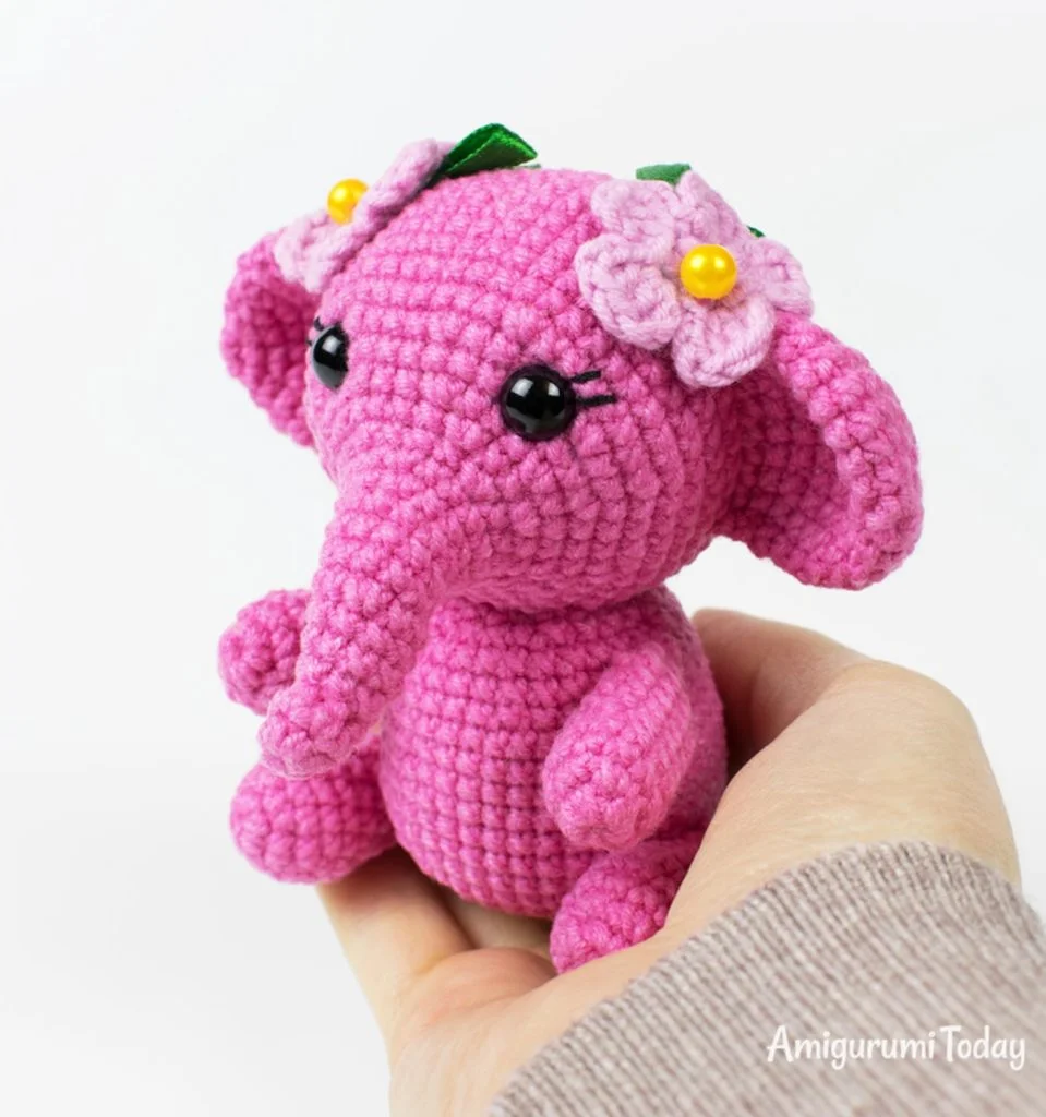 A hand holds a small pink crochet elephant toy decorated with pink and green fabric flowers.