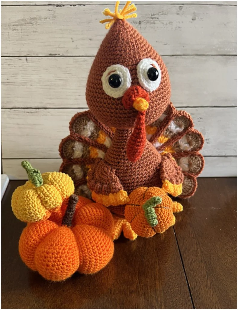 An amigurumi turkey with large eyes sits behind three crocheted pumpkins, all in autumn colors, on a wooden table with a white wood plank background.