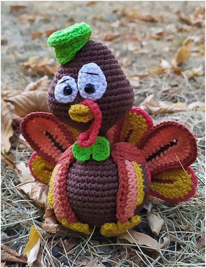 An amigurumi turkey doll with colorful wings and a green hat sits on dry grass with fallen leaves in the background.