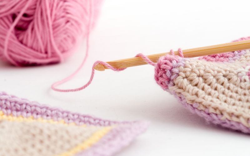 A crochet hook delicately works with pink yarn on a crochet piece, while a ball of luscious pink yarn sits patiently in the background, subtly inviting thoughts of what is gauge and its effects on crafting precision.