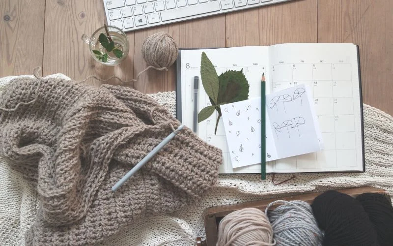 Knitting materials and a crochet piece, evoking thoughts on how to write crochet pattern tips, are displayed alongside a calendar, a plant, sketches, and yarn on a wooden table.