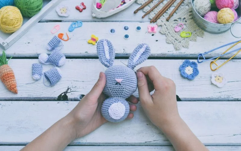 Hands delicately holding a crocheted bunny showcase the art of amigurumi, surrounded by vibrant crafting materials and the best yarn for achieving those perfect stitches on a rustic wooden table.