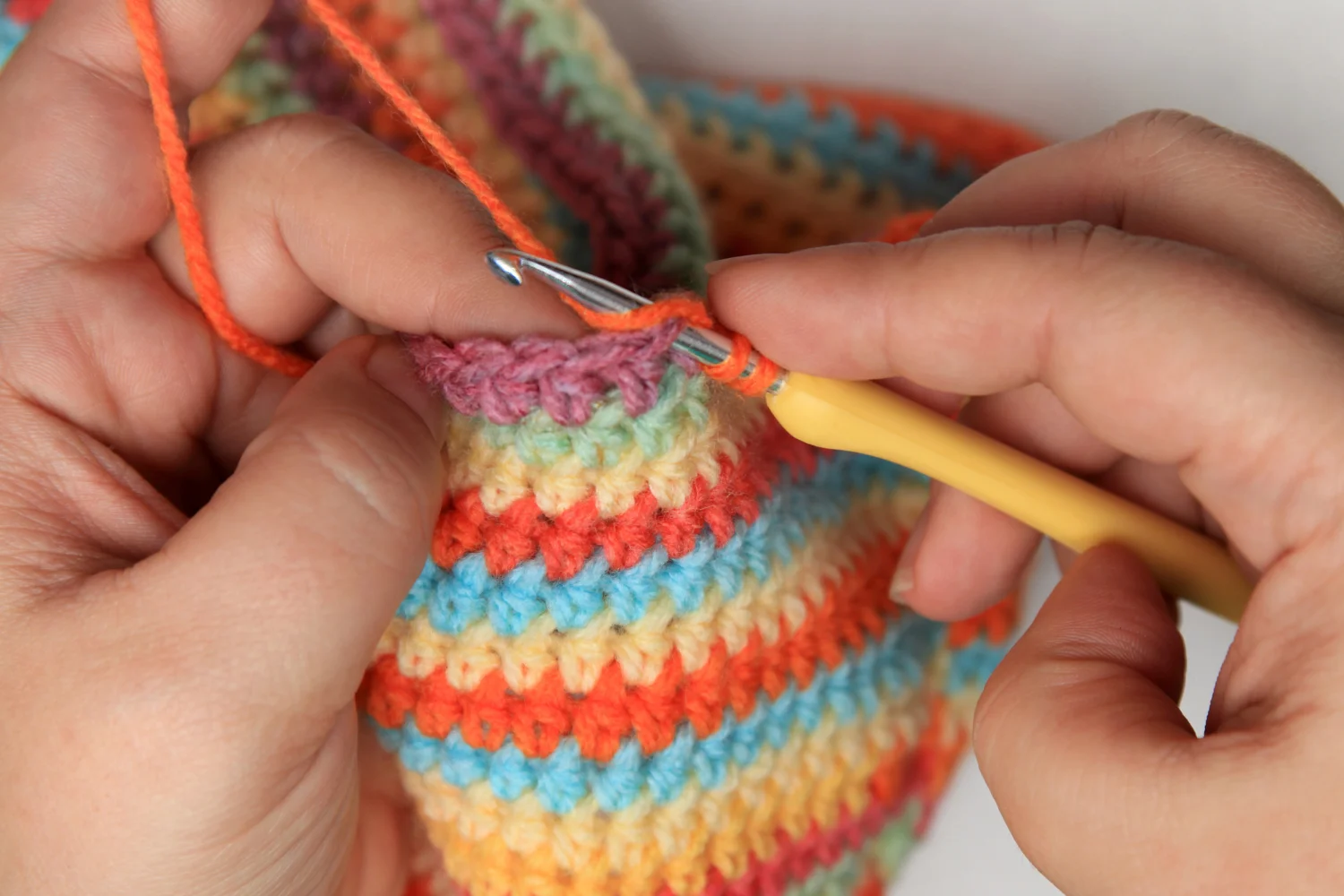 Hands crocheting a colorful striped fabric using an orange yarn and a yellow crochet hook, reminiscent of how to make a crochet afghan.