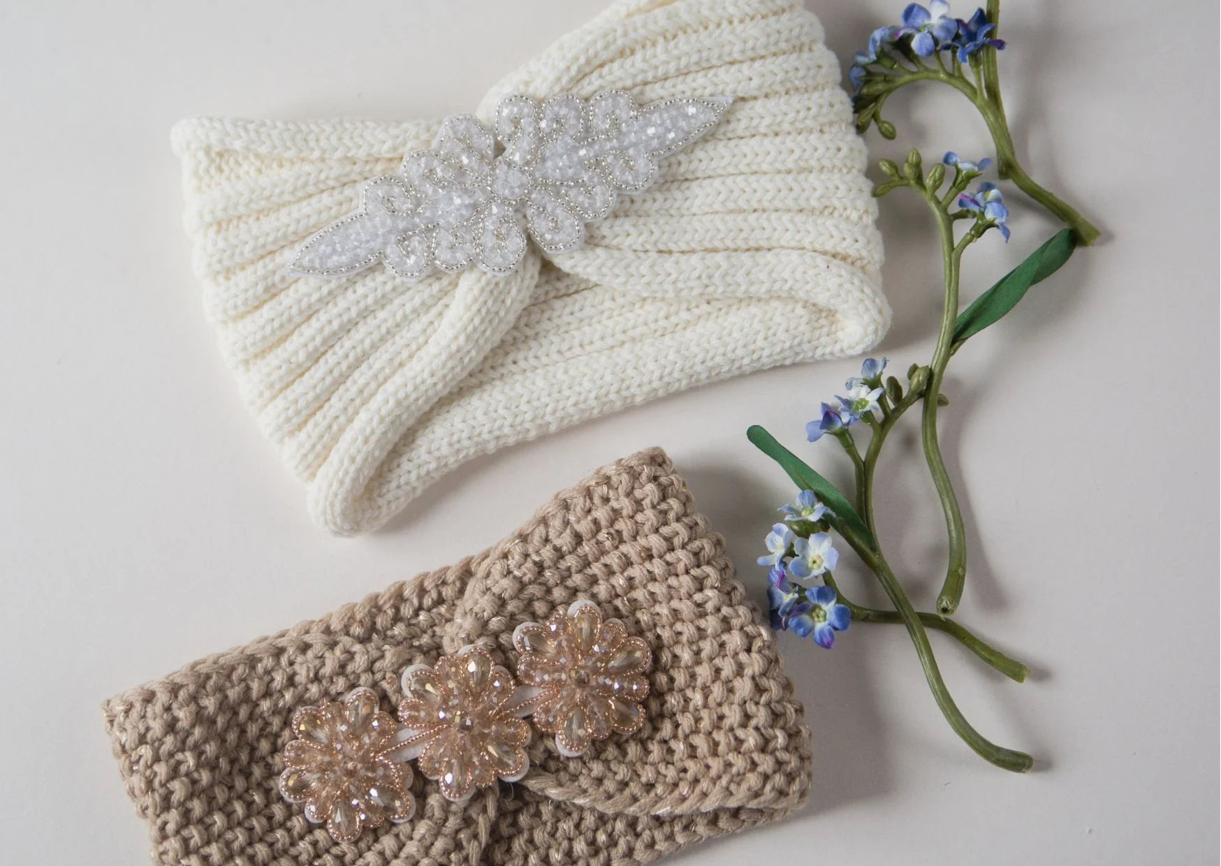 Two exquisite headbands, one a white crochet with a beaded design and the other a beige knitted piece adorned with floral embellishments, are beautifully displayed on a surface alongside sprigs of small blue flowers.