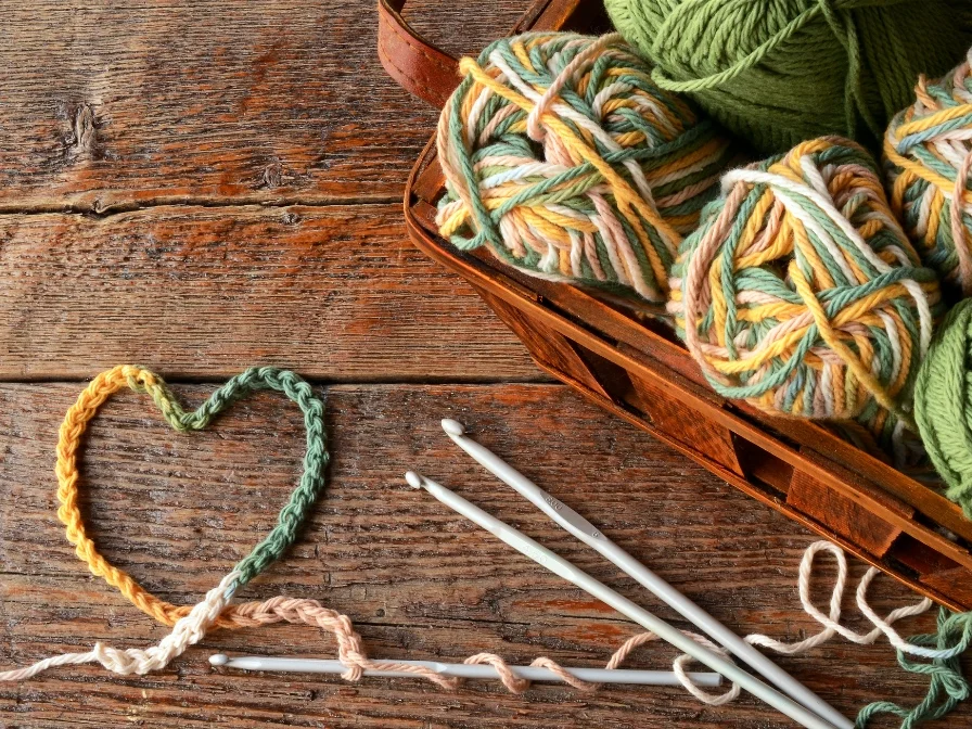 Yarn balls in a basket, crochet hooks, and a crocheted heart shape on a wooden surface hint at the start of a cozy project, perhaps a charming crochet sweater ready to bring warmth.